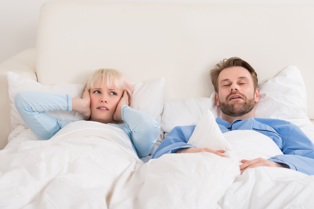 Couple laying in a white bed as the man sleeps and the woman covers her ears